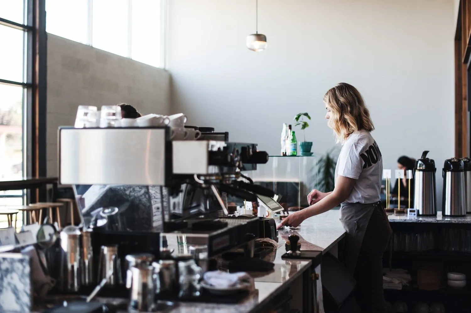 A barista at the espresso machine, a Hennepin Made glass pendant hanging above the bar