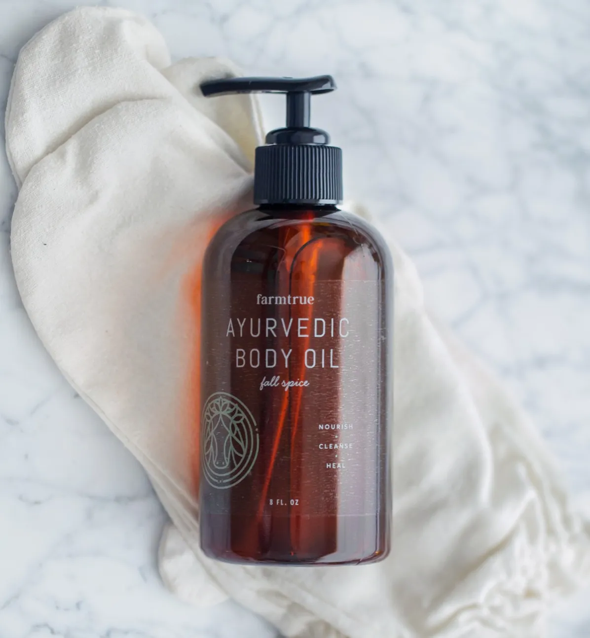 A FarmTrue Ayurvedic body oil pump bottle in amber glass, on linen