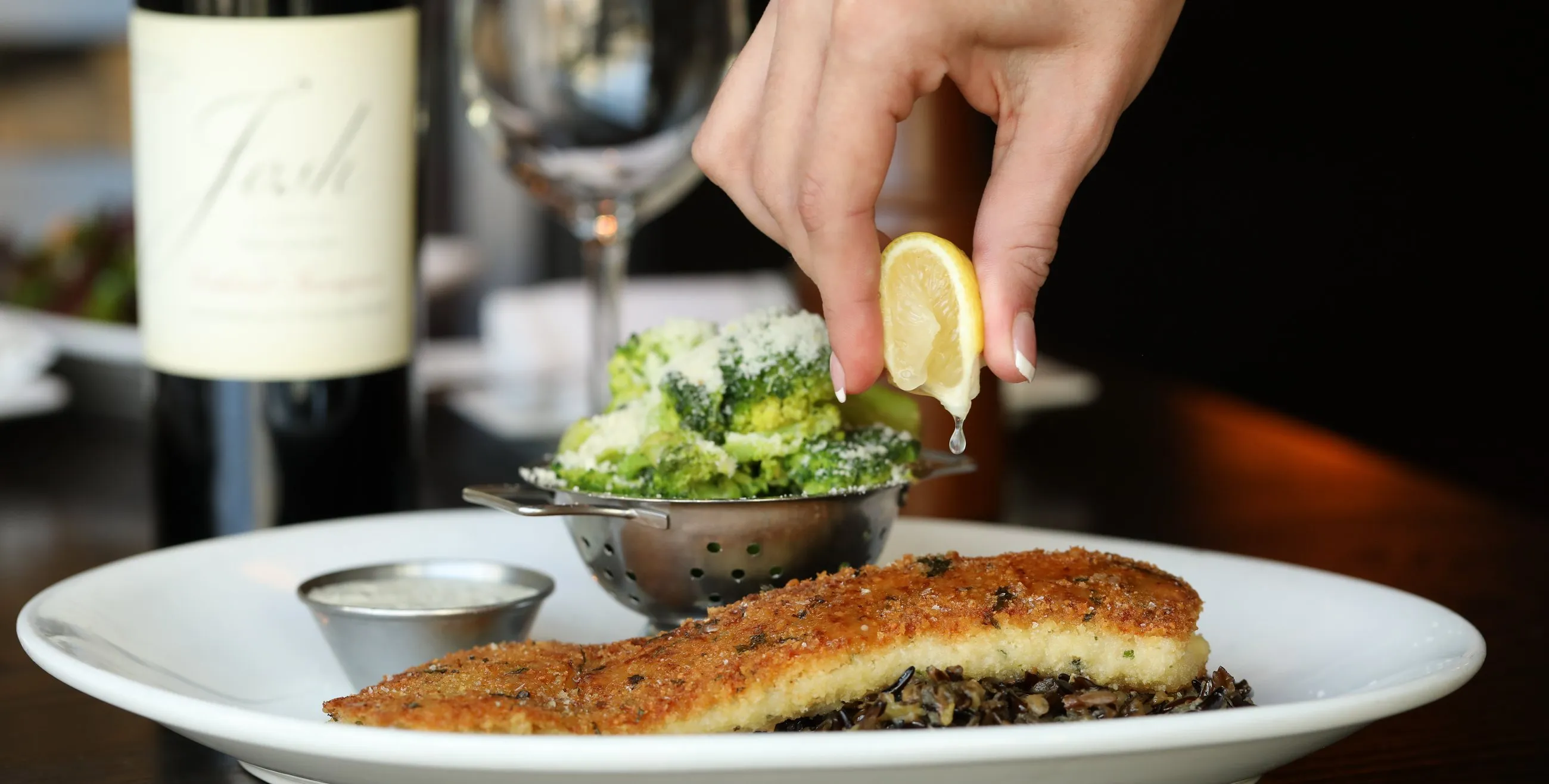 A Hazelwood fish entrée being finished with a squeeze of lemon, a glass of white wine behind