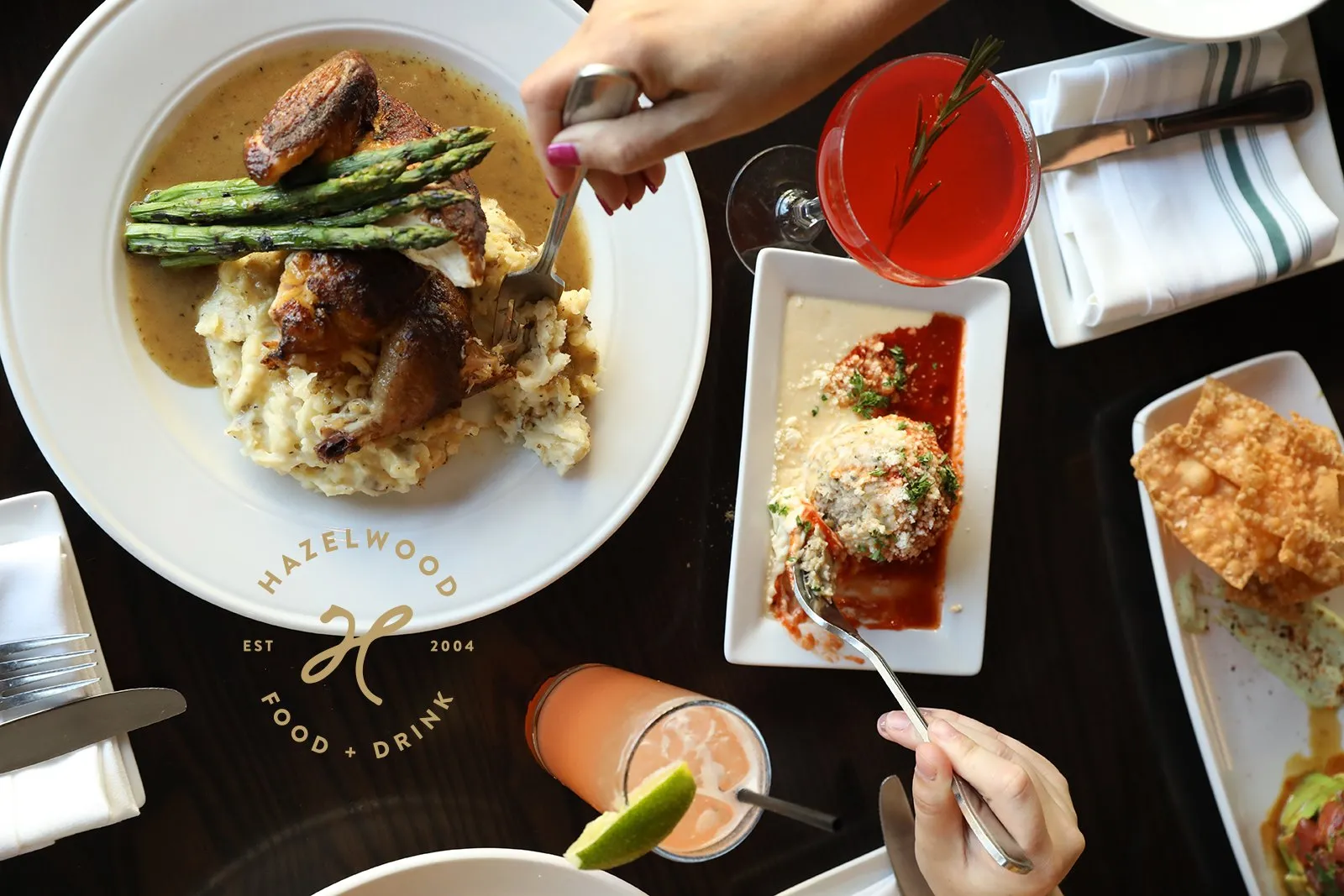 Overhead view of a Hazelwood table — rotisserie chicken with asparagus, a shared starter and cocktails — with the Hazelwood badge set on the dark wood