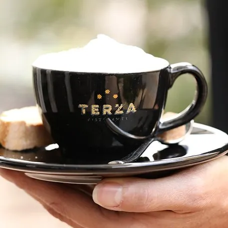 A black Terza cappuccino cup with the gold wordmark, held on its saucer
