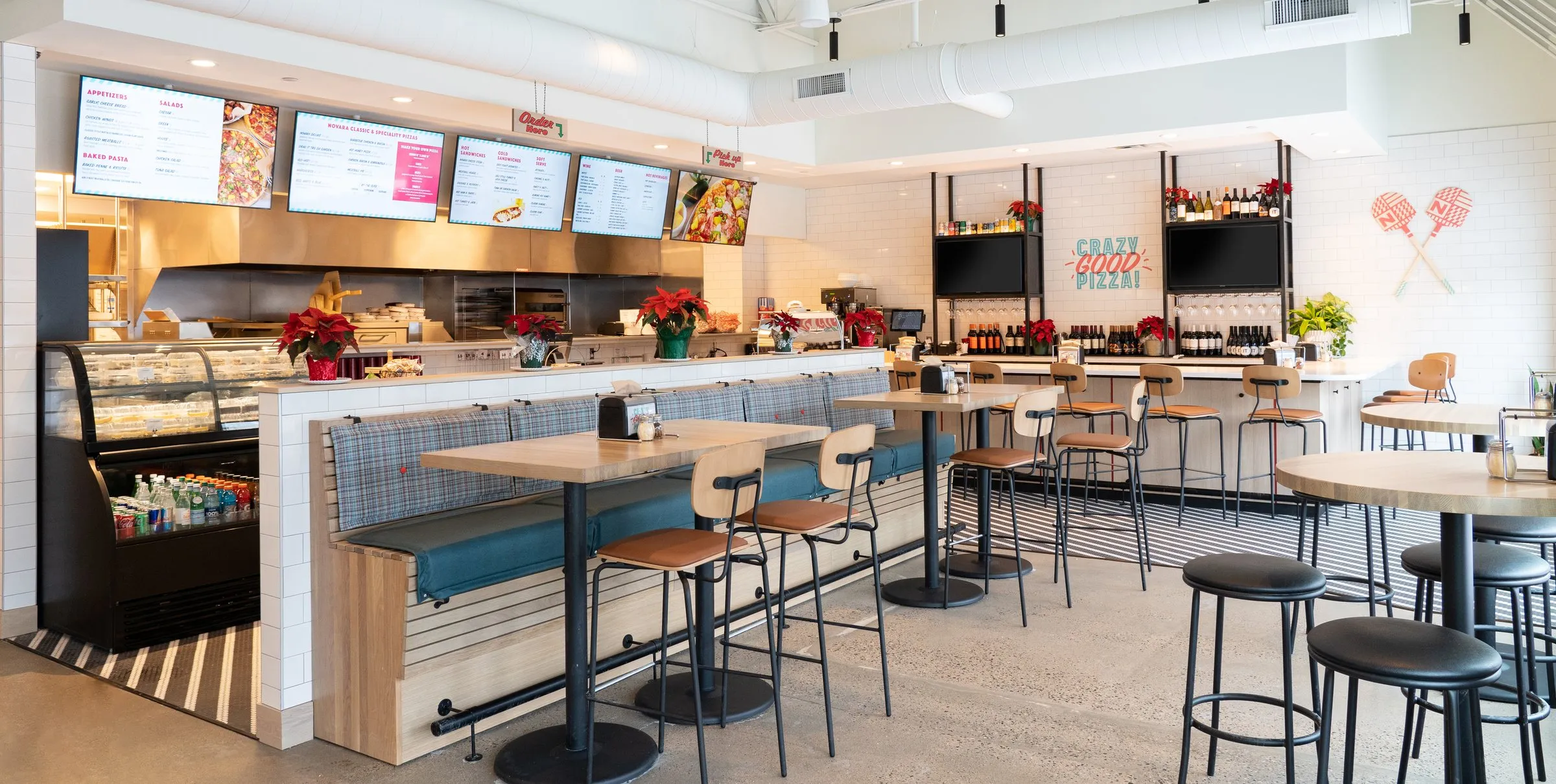 The Pizza Novara dining room: counter seating and high-tops facing the order counter, with four branded digital menu boards above the kitchen line