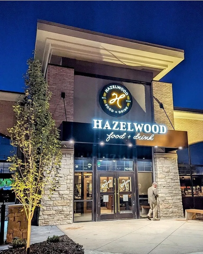The Hazelwood entrance at dusk: the lit H badge and Hazelwood food + drink sign above glass doors in a stone facade