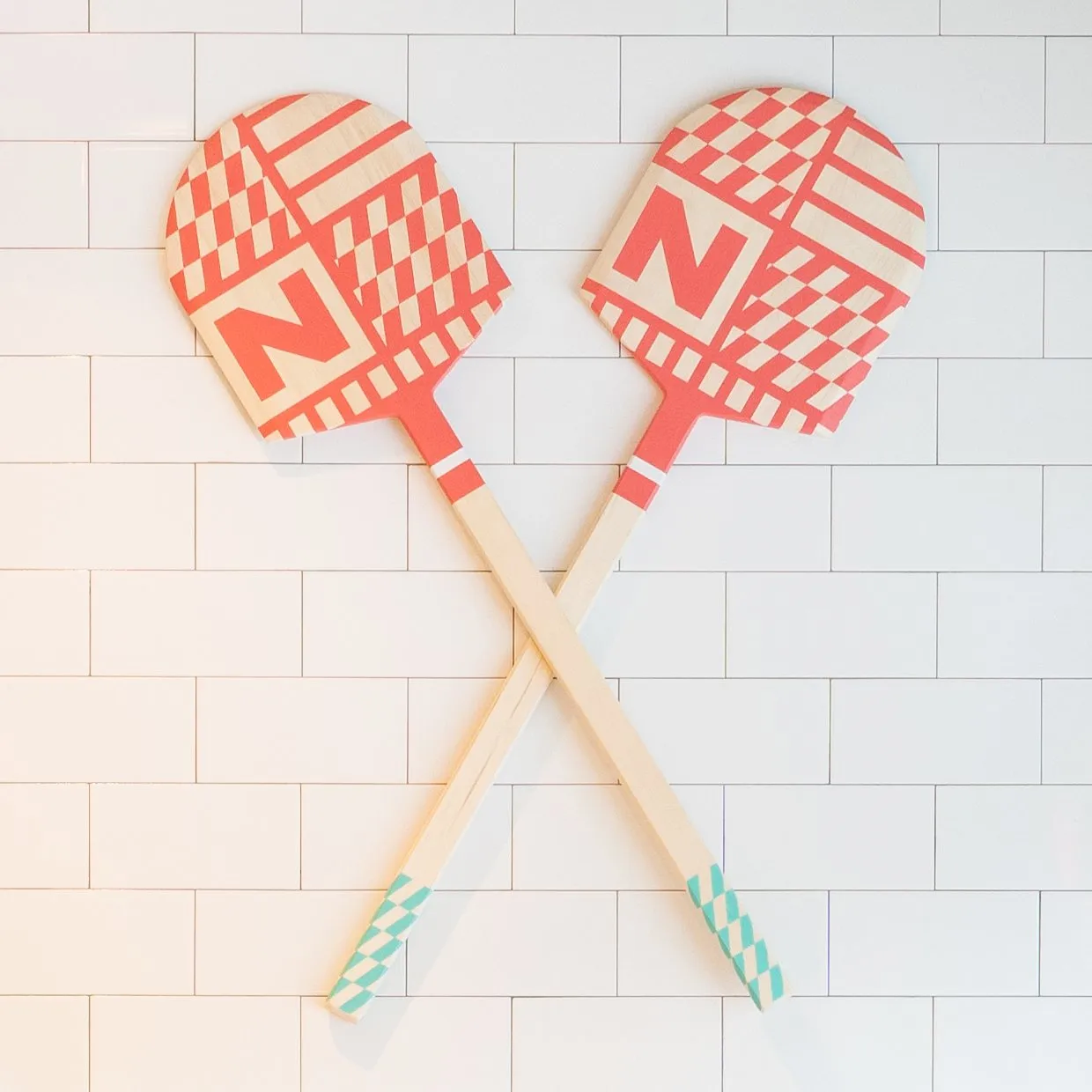 Two crossed pizza paddles printed with the Novara N and coral checkerboard, mounted on white subway tile