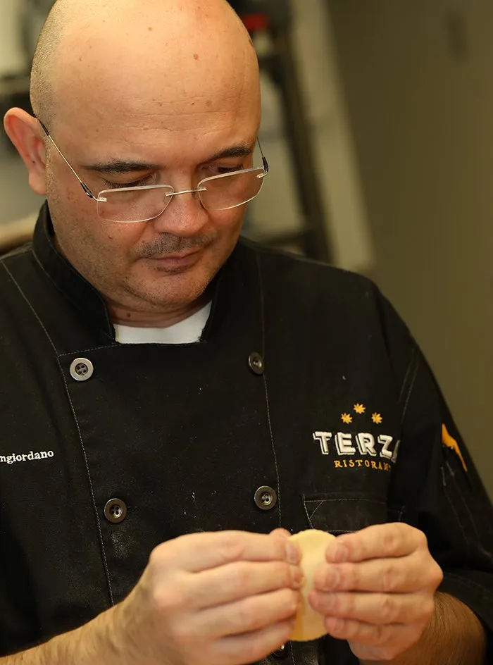 A chef in a Terza coat shaping pasta by hand, the wordmark embroidered on the chest