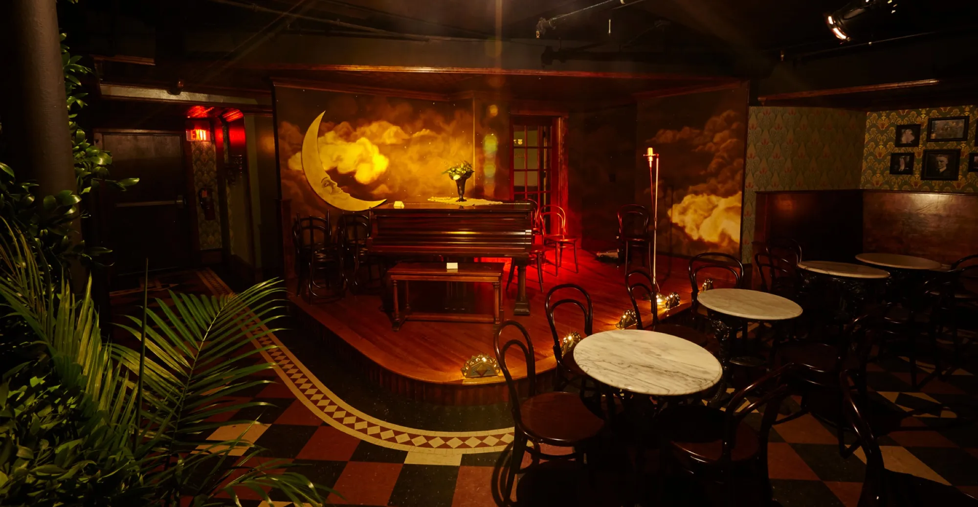 The Volstead’s Emporium room: a small stage under a painted moon and clouds, marble café tables and bentwood chairs on a checkered floor