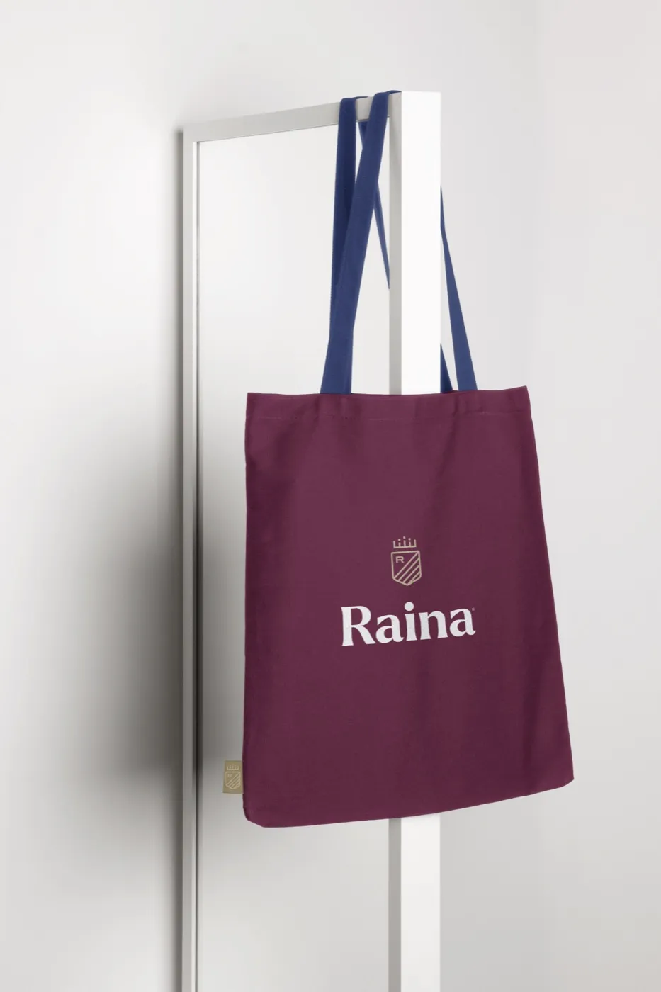 A plum Raina tote bag hanging from a hook, the crest and wordmark in cream