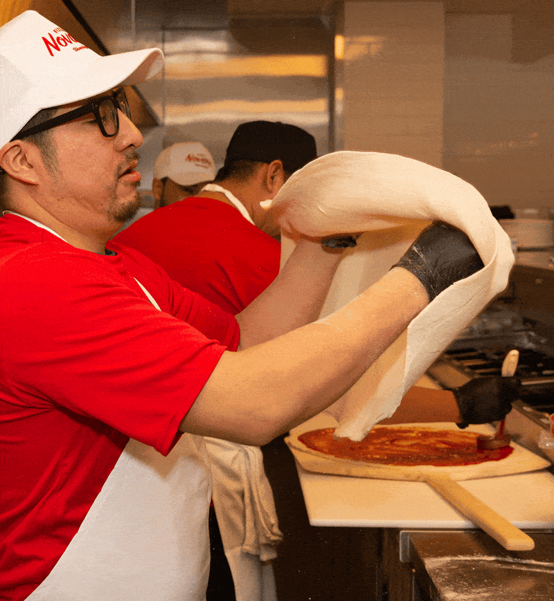 A Pizza Novara cook in a branded cap stretching and tossing dough above the make line